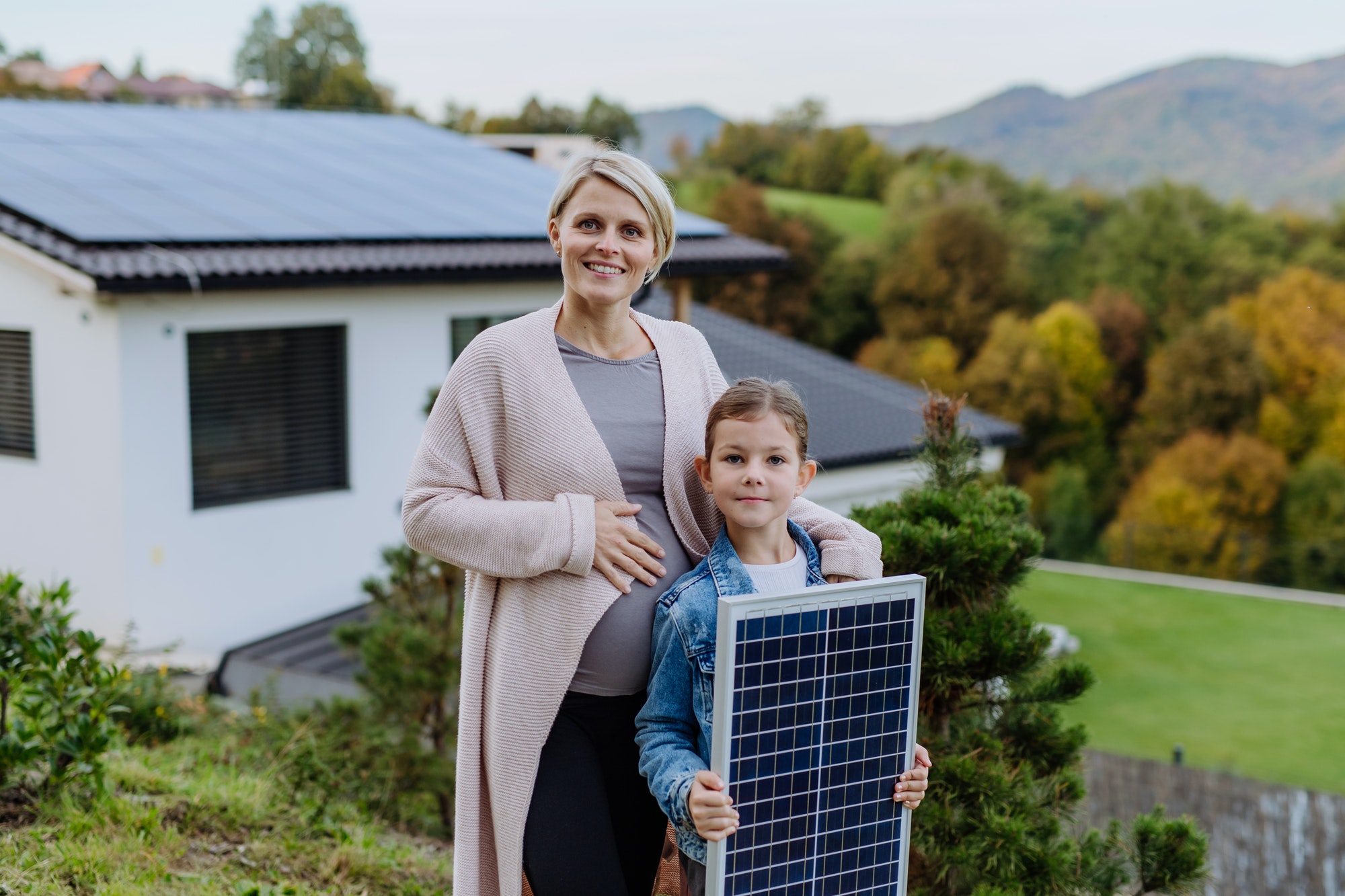 Mother with her daughter near their house with solar panels. Alternative energy, saving